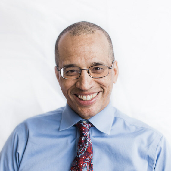 Reed Colfax is smiling. He has short-cropped black hair, is wearing glasses, a red and blue paisley tie and a blue shirt.