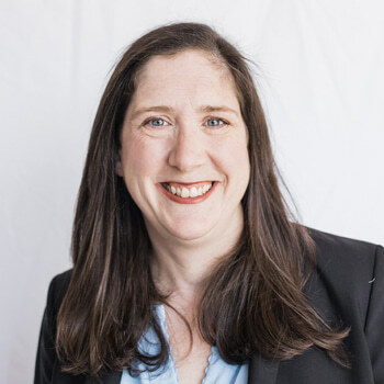 Jennifer Klar is a white woman in her 40s with long brown hair and blue eyes. She is wearing a light blue shirt and a black blazer and smiling.