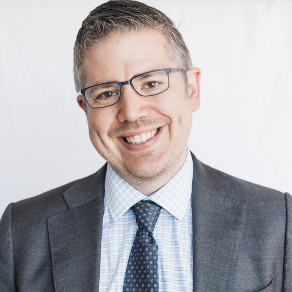 Eric Sublett is a white man in his 40s with short, gray hair. He is wearing a gray suit with a patterned tie and glasses. He is smiling.