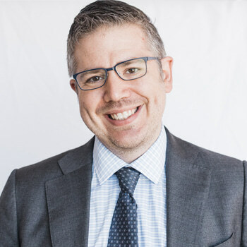 Eric Sublett is a white man in his 40s with short, gray hair. He is wearing a gray suit with a patterned tie and glasses. He is smiling.