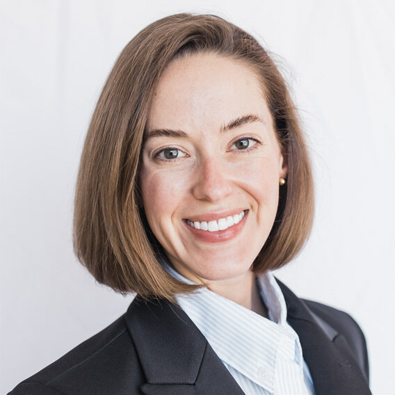 Danielle Root is a white woman in her 30s with brown hair and brown eyes. She is wearing a blue shirt and black jacket, and she is smiling.