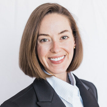 Danielle Root is a white woman in her 30s with brown hair and brown eyes. She is wearing a blue shirt and black jacket, and she is smiling.