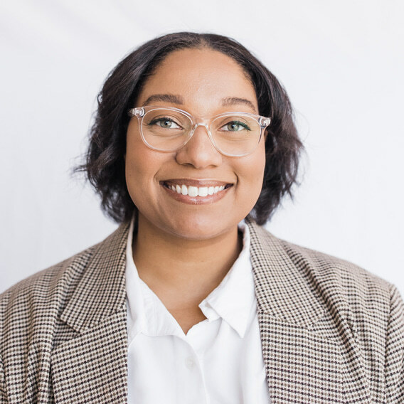 Imani Cherry (she/her) is a Black woman in her 30s with short black hair and glasses. She is wearing a brown checkered blazer and white collared shirt and is smiling.