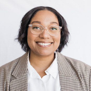 Imani Cherry (she/her) is a Black woman in her 30s with short black hair and glasses. She is wearing a brown checkered blazer and white collared shirt and is smiling.