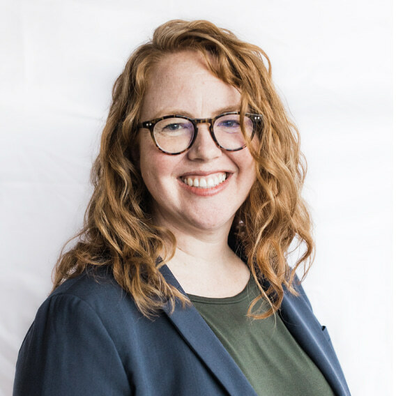 Alexa Milton is a roughly 40-year-old white woman with wavy red hair and glasses. She is wearing a dark green shirt and navy suit jacket and is smiling.  