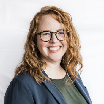 Alexa Milton is a roughly 40-year-old white woman with wavy red hair and glasses. She is wearing a dark green shirt and navy suit jacket and is smiling.  