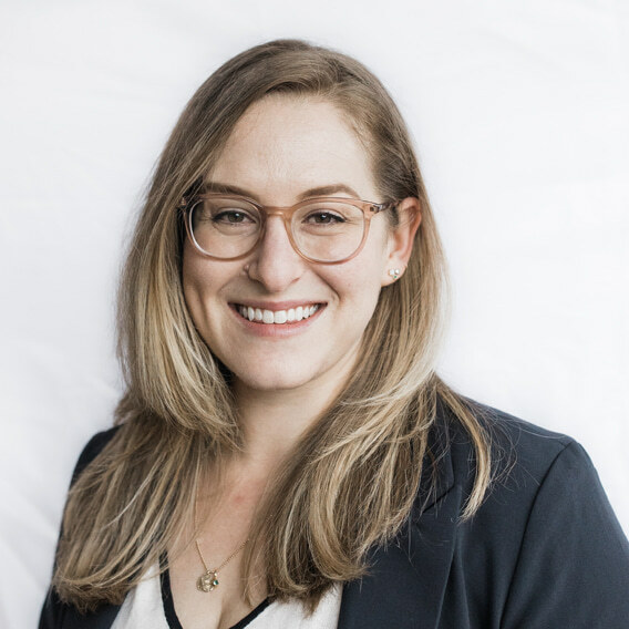 Sacha Markano-Stark is a white woman in her 30s with long blonde hair and glasses with rose-colored frames.  She is wearing a white shirt with black trim, a black suit jacket, and a necklace, and is smiling.