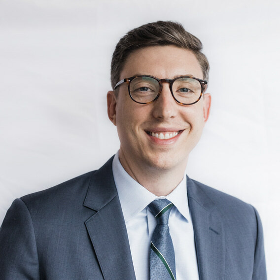 Zachary Best is a white man in his late 30s with brown hair and blue eyes.  He is wearing a blue suit and glasses and is smiling