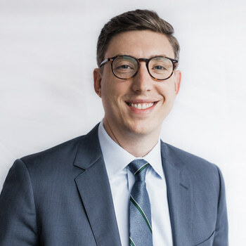 Zachary Best is a white man in his late 30s with brown hair and blue eyes.  He is wearing a blue suit and glasses and is smiling