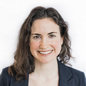 Brenna Kearns is a white woman in her 30s with brown hair. She is wearing a dark suit and metallic earrings and is smiling.