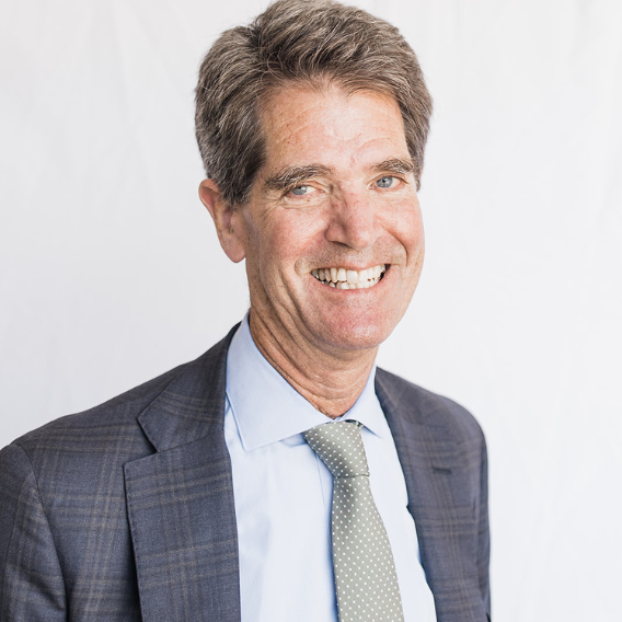 John Relman is a white man in his late sixties.  He is wearing a blue suit and a green tie and is smiling. 