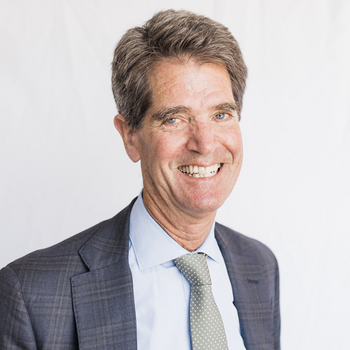 John Relman is a white man in his late sixties.  He is wearing a blue suit and a green tie and is smiling. 