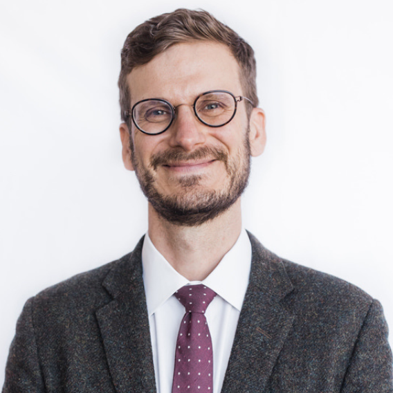 Ted Olds is a middle-aged white man with round glasses and a short beard. He is wearing a tweed sports coat and a marron tie with white polka dots and is smiling.