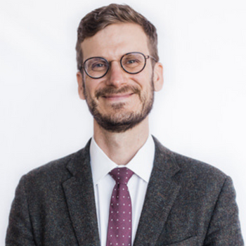 Ted Olds is a middle-aged white man with round glasses and a short beard. He is wearing a tweed sports coat and a marron tie with white polka dots and is smiling.