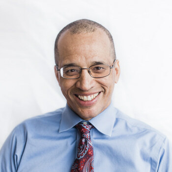 Reed Colfax is smiling. He has short-cropped black hair, is wearing glasses, a red and blue paisley tie and a blue shirt.