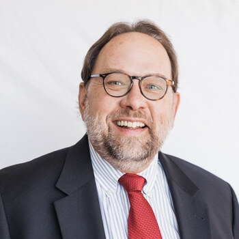 Michael Allen is a white man in his mid-60s with a beard and glasses. He is wearing a dark blue suit and a red tie and is smiling.