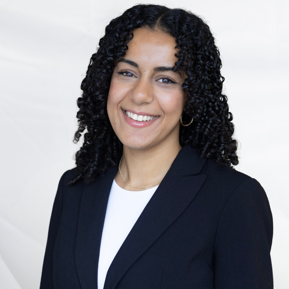 Miriam Farah is a Middle Eastern woman in her early twenties. She has black curly hair and dark brown eyes. She is wearing a blue suit and is smiling.