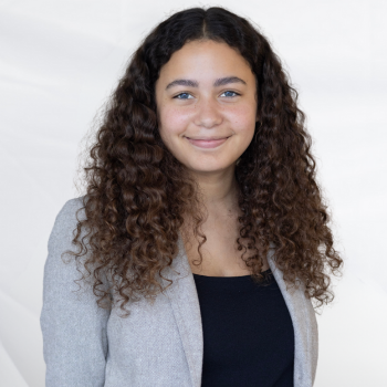 Jazmin Trenco is a Latina woman in her 20s with brown curly hair. She is wearing a light gray blazer and is smiling.