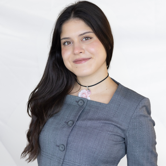 Esmeralda Hermosillo is a Latina woman in her early 20s with brown straight hair, she is wearing a grey suit with a rose pendant.