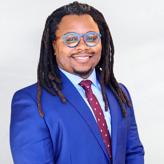 Emahunn Raheem Ali Campbell is a Black man in his late-30s with locs and glasses. He is wearing a royal blue suit and a maroon tie and is smiling.