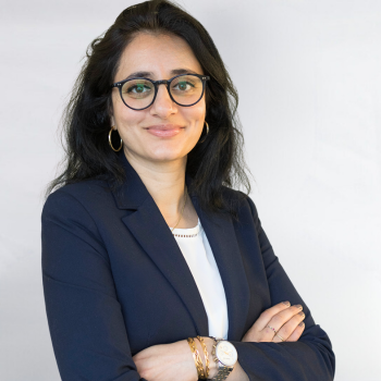 Ellora Israni is a South Asian woman in her 30s. She is wearing a blue jacket and glasses and smiling.