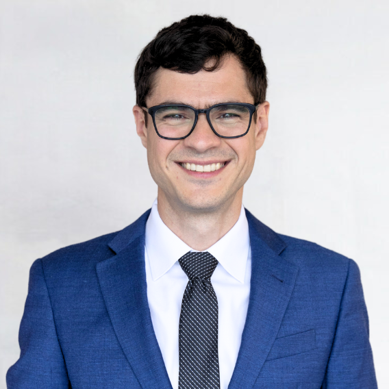 Robert Hunter is a man in his mid-30s with dark brown hair and glasses. He is wearing a blue suit and black tie and is smiling.