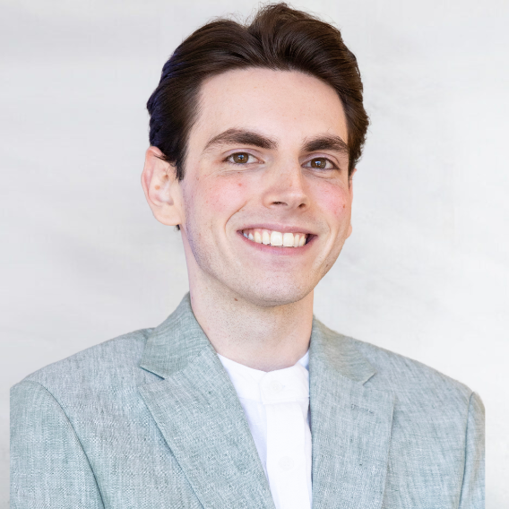 Jake Hogan is a white man in his mid twenties with brown hair and eyes. He is wearing a Green Suit and Smiling.