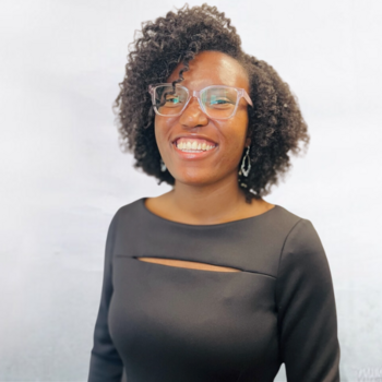 Taylor Gaskins is a Black (African American) woman in her 20’s with brown eyes and glasses. She is wearing a black dress and standing against a white backdrop, smiling.