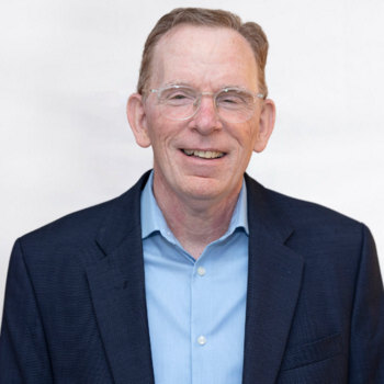 Tim Lambert is a white man in his mid-60s with glasses. He is wearing a dark blue jacket and light blue shirt and is smiling.