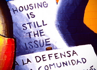 A wall mural with phrases including "Housing is Still the Issue" and "Defensa de la Comunidad"