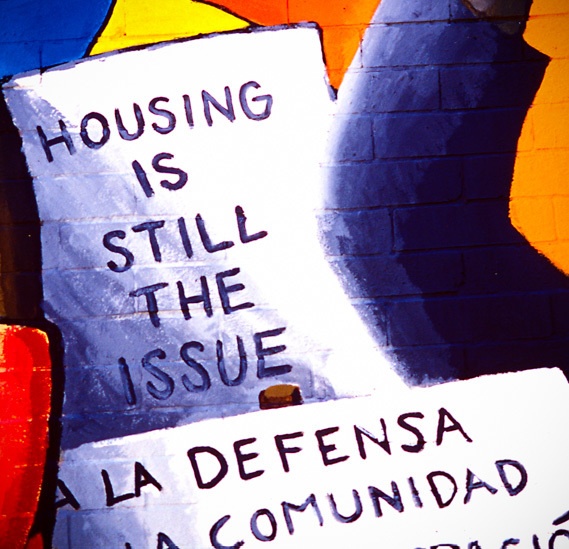 A mural with bold, black text reading 'Housing is still the issue' painted on a wall. Below, partially visible Spanish text reads 'A la defensa ... la comunidad,' suggesting a message about community defense.