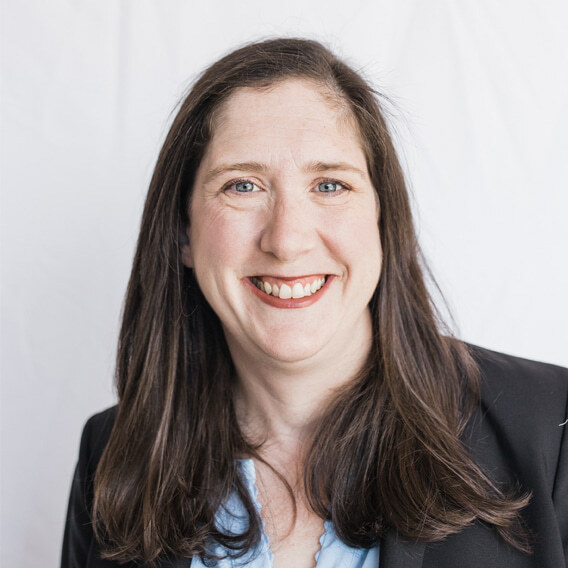 Jennifer Klar is a white woman in her 40s with long brown hair and blue eyes. She is wearing a light blue shirt and a black blazer and smiling.